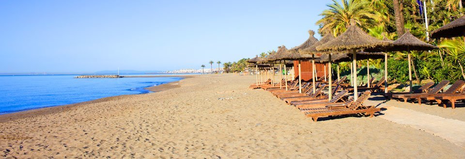 Billedet viser en solrig strand med parasoller af strå og liggestole, der vender mod et klart blåt hav.