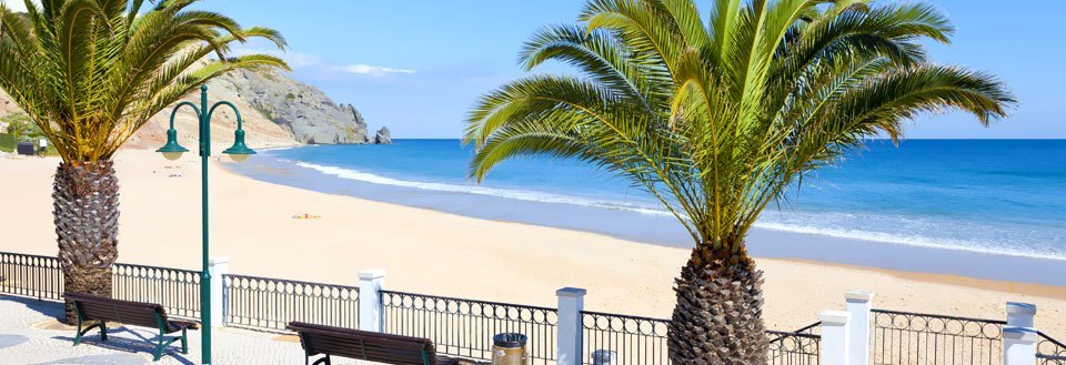 En solrig strandpromenade med palmetræer, bænke og udsigt til havet og en bjergklippe i det fjerne.