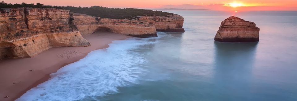 Solnedgang med bløde farver over en strand med klippeformationer i havet.