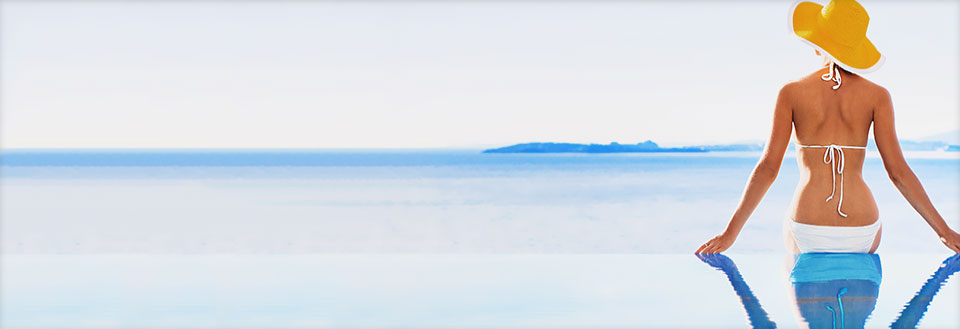 Kvinde med solhat står ved en pool, ser ud over et roligt hav mod en horisont med øer.
