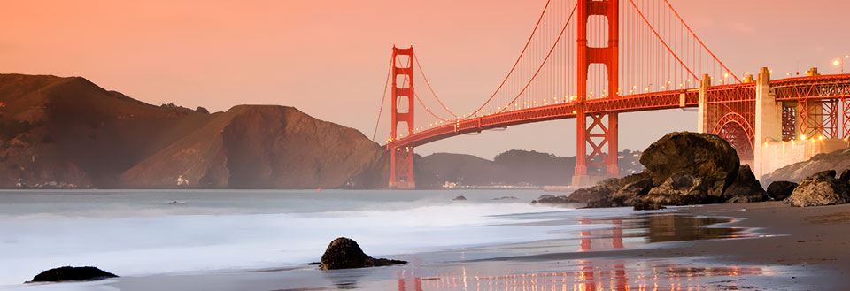 Solnedgang bag Golden Gate Bridge med roligt vand og strand foran.