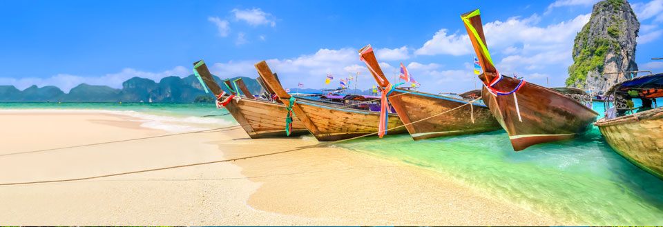 Traditionelle træbåde ved en tropisk strand med klart vand og dramatiske klippeformationer i baggrunden.