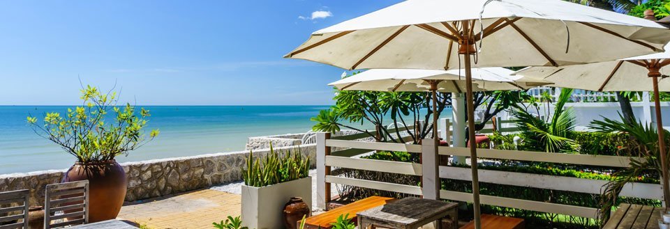 En solrig terrasse med parasoller og havemøbler ud til en rolig azurblå havfront og klar himmel.