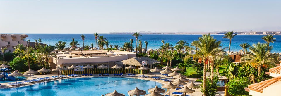 Billedet viser et ferieresort i  med poolområde, parasoller og palmer med havet i baggrunden.