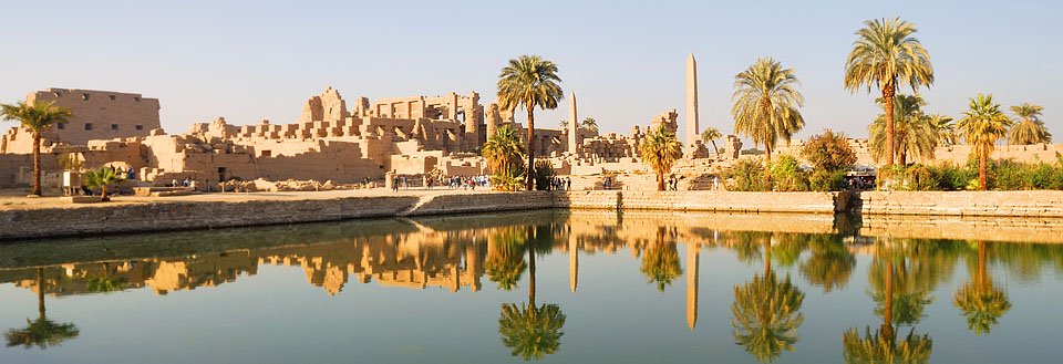 Billedet viser gamle ruiner reflekteret i vandet med palmetræer og en klar himmel.