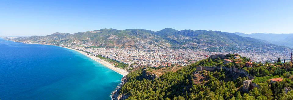 Panoramaudsigt over Antalya med blåt vand, strande og grønklædte bjerge i baggrunden.