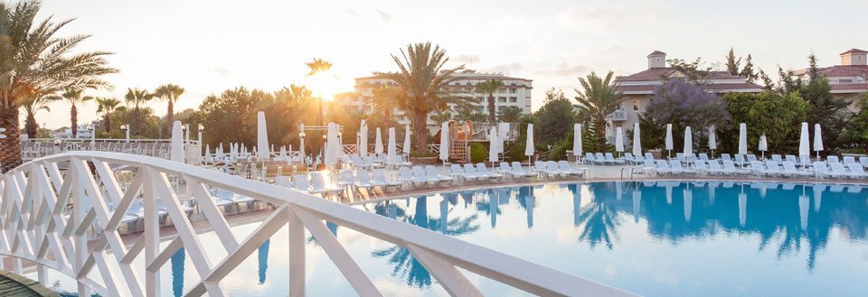 Solnedgang over rolige resort i Side med swimmingpool, hvidlakerede liggestole og palmer.