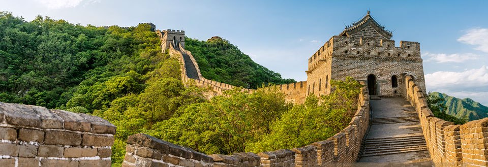 Panoramabillede af Den Kinesiske Mur strækker sig over frodige grønne bjerglandskaber under en blå himmel.