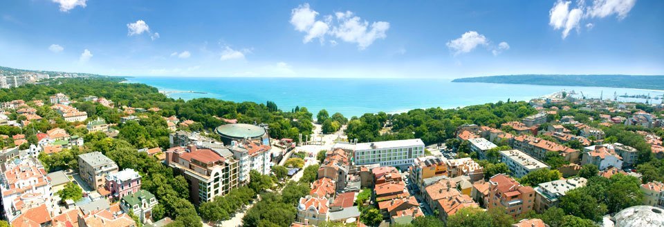 Panoramaudsigt over en Varna med tætte boliger, omgivet af træer og med et azurblåt hav i baggrunden.