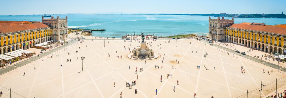 Panoramaudsigt over en rummelig, åben plads i Lissabon med gående mennesker, flankeret af klassiske bygninger og et hav i baggrunden.