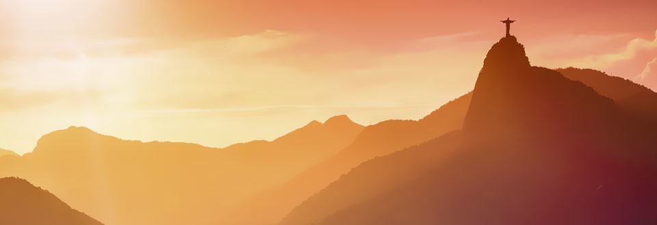 Solnedgang bag bjerge med Kristusstatuen på toppen i silhuet mod en glødende himmel.