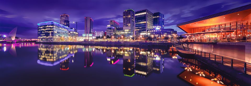 Manchesters skyline om aftenen med lysende bygninger og deres spejlinger i vandet.