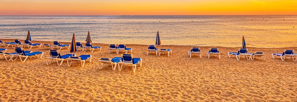 Billede af tomme liggestole og parasoller på en sandstrand med rolig hav i baggrunden ved solnedgang.