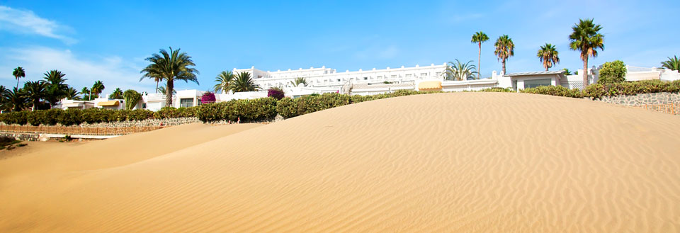 Ørkenlignende sandklit foran en række hvide bygninger flankeret af palmer under en klar blå himmel.