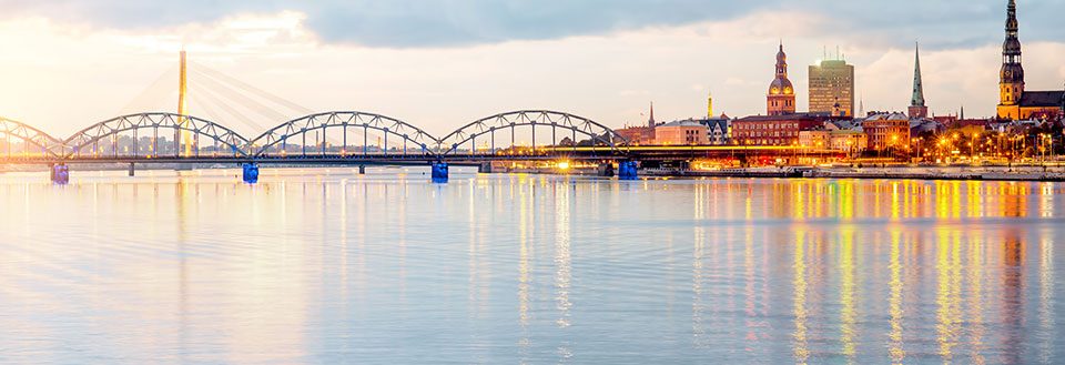 Billede af Riga ved floden i skumringen med lys der reflekterer i vandet og en bro i baggrunden.