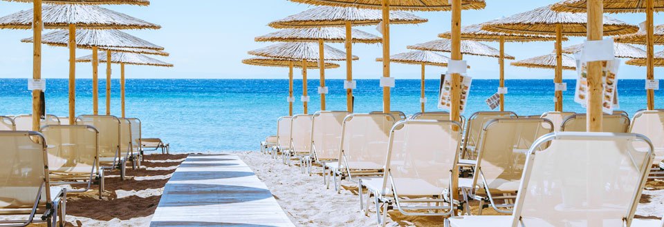En solrig strand med parasoller af strå og rækker af tomme liggestole, der vender mod det blå hav.