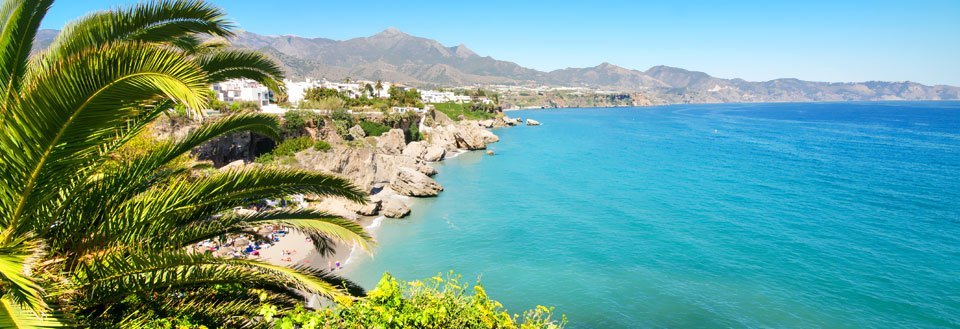 Panoramaudsigt over en frodig kystlinje med palmer, klipper og en klar blå hav. Bjerge ses i baggrunden.