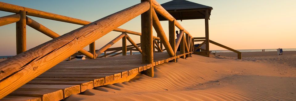 En træbro fører gennem sanddynene til en strand ved solnedgang med en klar himmel.