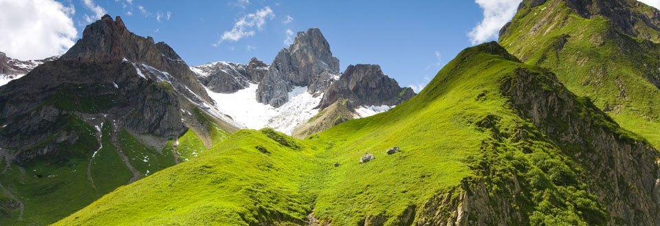 Frodig grøn dal med dramatiske bjergtoppe og snedækkede tinder i baggrunden under en klar blå himmel.