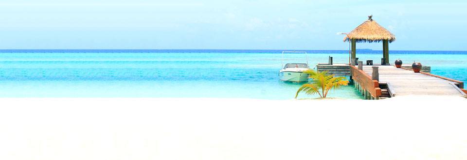 Et idyllisk strandbillede med hvidt sand, en træbro der fører ud til en hytte med stråtag og en båd.