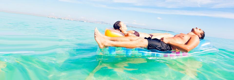 To personer slapper af på en oppustelig madras i det krystalklare havvand under en blå himmel.