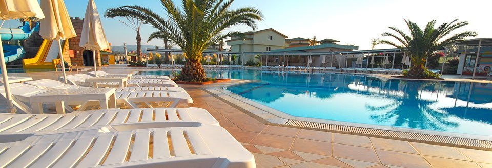 Et feriested med swimmingpool, solsenge og palmetræer i solnedgangens lys.