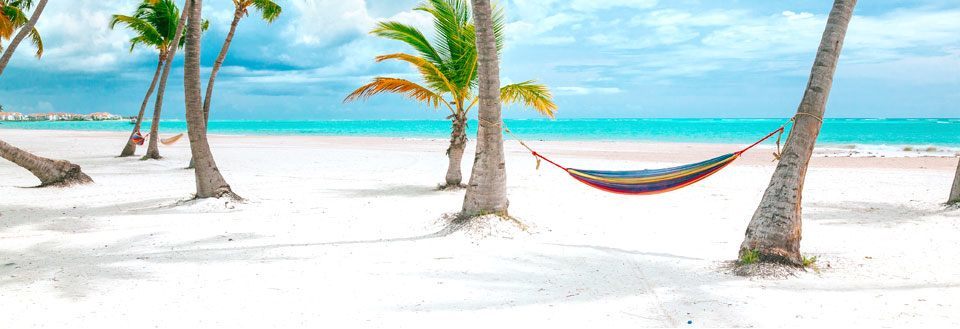 En hængekøje mellem palmer på en hvid sandstrand med klart blå himmel og hav i baggrunden.