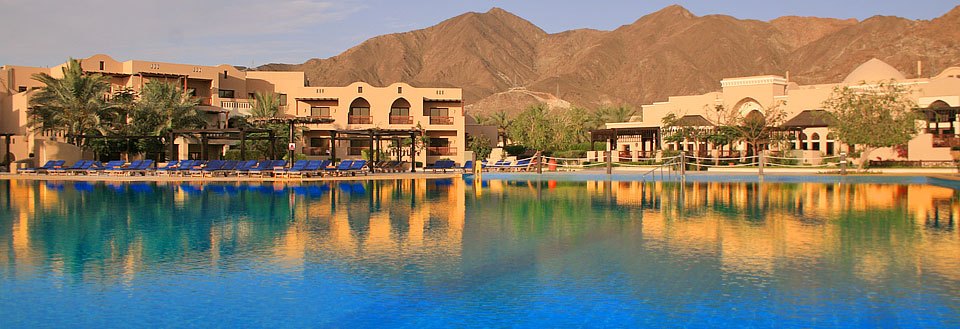 Resort med swimmingpool foran bjerge, liggestole og parasoller, klart blåt vand og solskin.