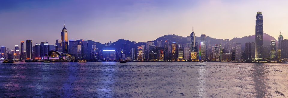 Skumring over Hong Kongs skyline med højhuse og oplyste bygninger, reflekteret i vandet foran.