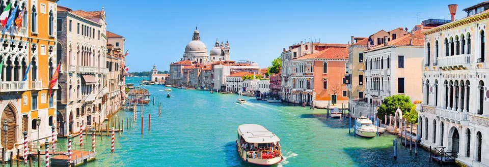 Billedet viser en klar dag i Venedig med historiske bygninger langs Canal Grande og en båd på vandet.