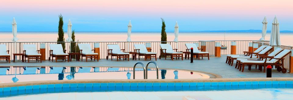 Et fredfyldt poolområde ved solnedgang med liggestole og lukkede parasoller. Havudsigt i baggrunden.