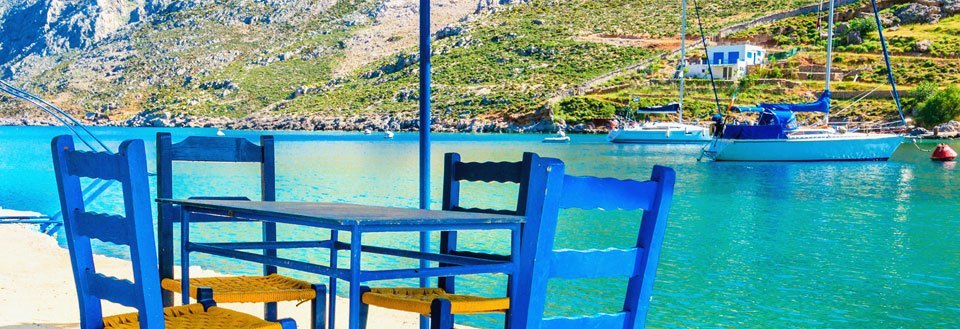 Et idyllisk strandbillede med blå stole og bord foran en klar blå bugt med både og bjerge i baggrunden.