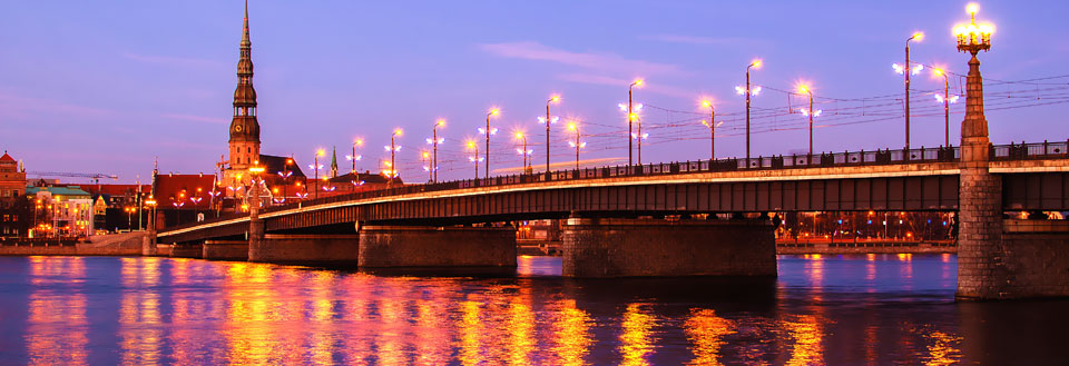 Aftenbillede af en oplyst bro over en flod, med bygninger og et tårn i baggrunden.