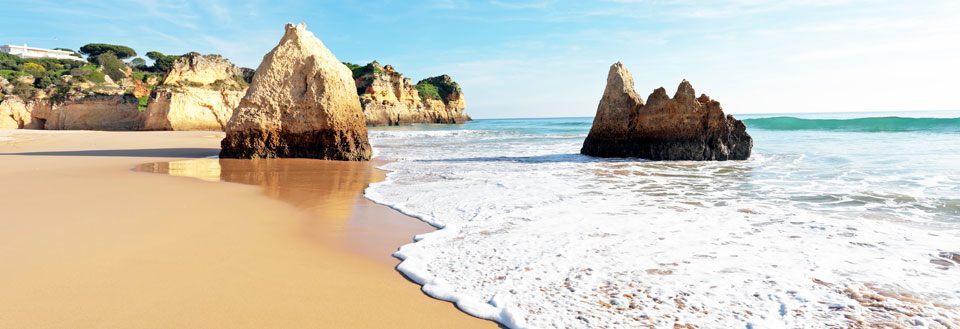 Billede af en fredelig strand med gyldent sand og store klippeformationer, og bølgerne skvulper ind.