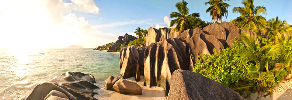 Et tropisk strandpanorama med store klippeformationer, palmer, sand og et glitrende hav.
