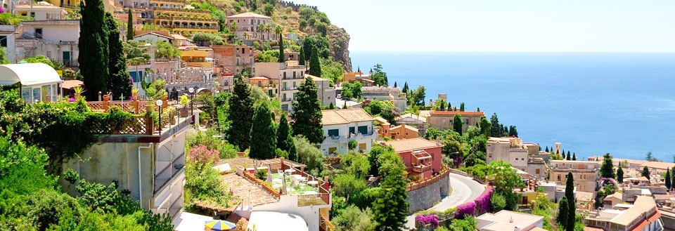 Billede af en malerisk kystby med farverige huse og frodig vegetationsklædt terræn mod en klar blå himmel.