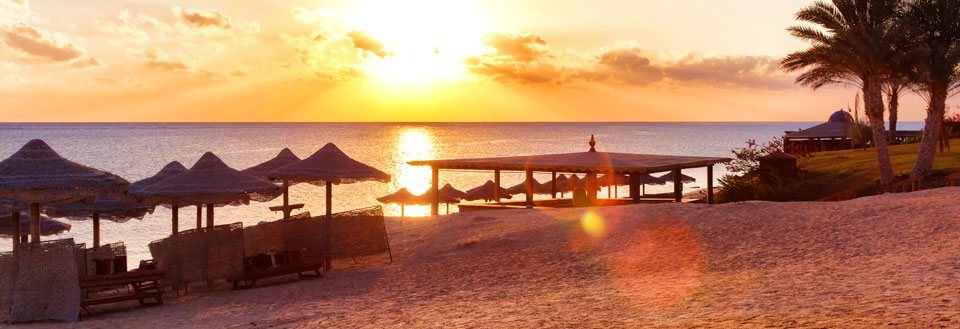 Solnedgang på en strand med stråparasoller og liggestole. Havet spejler solens stråler, mens himlen gløder varmt.