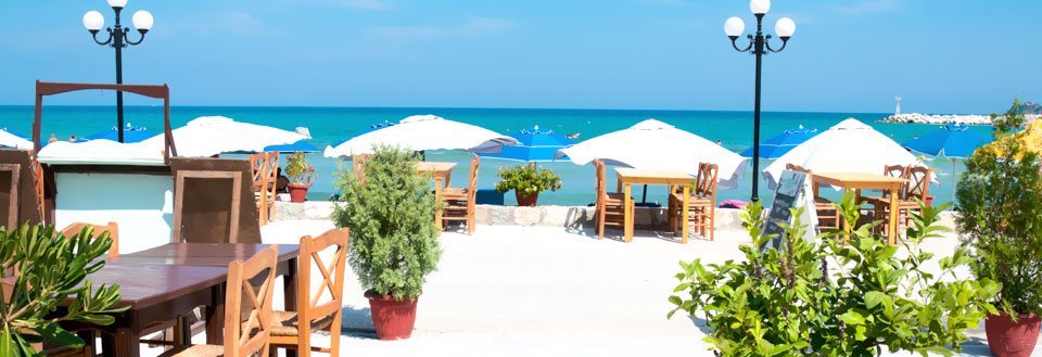En idyllisk strandcafé med træborde og -stole under hvide parasoller. Havet glitrer i baggrunden.