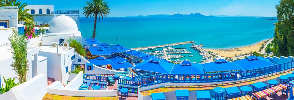 Panoramaudsigt over en solrig kystby med hvidkalkede bygninger, blå parasoller og et azurblåt hav.