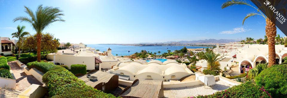 Panoramaudsigt over et luksuriøst feriested med hvidkalkede bygninger, palmer og havudsigt.