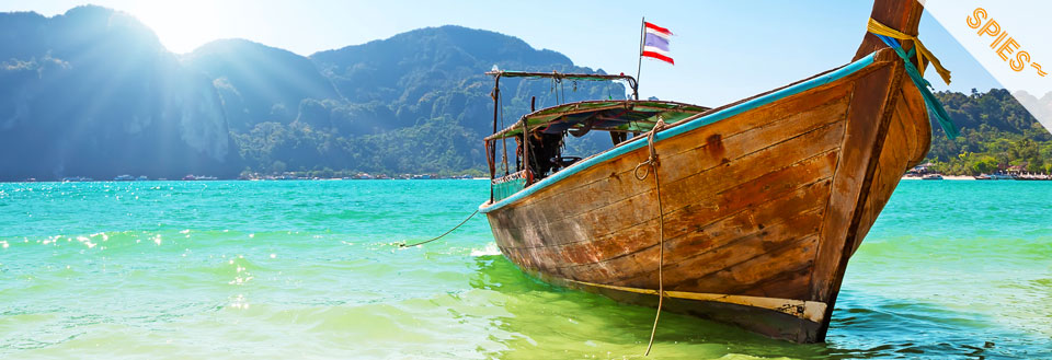 En traditionel båd flyder nær kysten med klare blågrønne vande og bjerge i baggrunden.