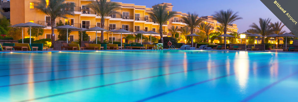 Et luksuriøst resort med swimmingpool i skumringen. Liggestole og parasoller står ved poolkanten.
