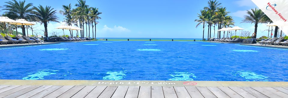 En luksuriøs udendørs swimmingpool med klart blåt vand omgivet af palmer og liggestole under en klar himmel.