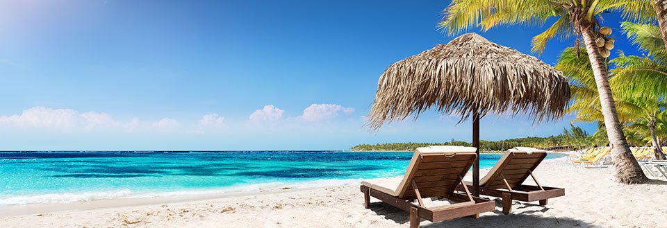 To solsenge under en stråparasol på en hvid sandstrand med klart blåt hav og palmer.