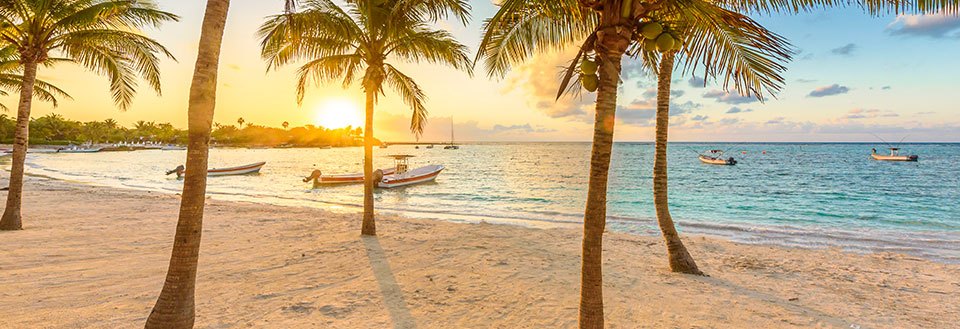 Solnedgang ved en tropisk strand med palmer og små både i havet.