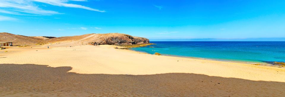 Billede af en solrig strand med gyldent sand, krystalklart blåt hav og en stenet klippe i baggrunden.