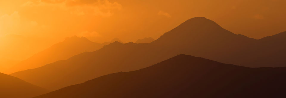 Billedet viser silhuetten af bjergkæder mod en gylden solnedgang med varme farver.