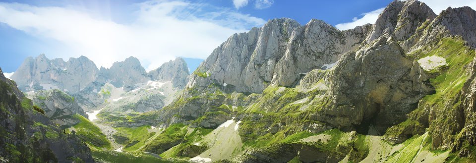Billedet viser en bjergkæde med grønne dale, spidse tinder og blå himmel.