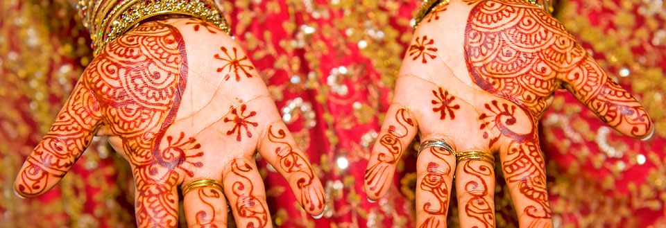 Hænder dekoreret med detaljeret henna kunst foran en rød gylden baggrund.