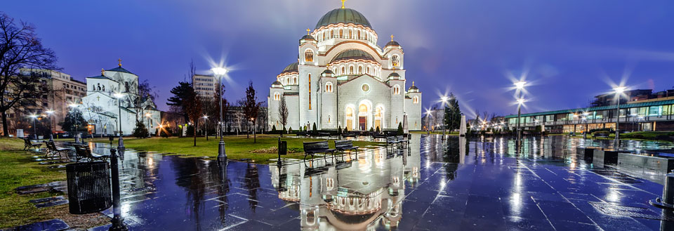 Billedet viser en oplyst kirke om aftenen med vådt fortov, der spejler bygningerne og lysene.
